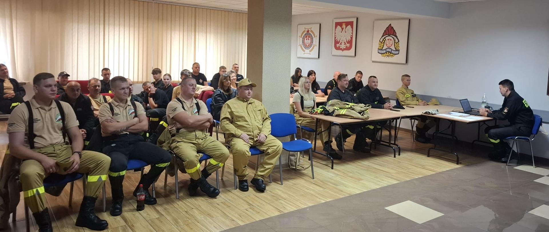 Grupa strażaków ochotników uczestniczy w szkoleniu podstawowym, siedząc w sali wykładowej. Ubrani są w umundurowanie bojowe w kolorze piaskowym oraz czarnym. Prowadzący, strażak w ciemnym stroju z napisem "PSP", siedzi przy stole z laptopem i prowadzi zajęcia teoretyczne. W tle na ścianie wiszą godła i emblematy związane ze strażą pożarną, w tym godło Polski. Uczestnicy są skupieni, słuchają z uwagą.