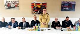 Strażak Państwowej Straży Pożarnej przemawia podczas spotkania obok siedzą druhowie ochotniczych straży pożarnych.