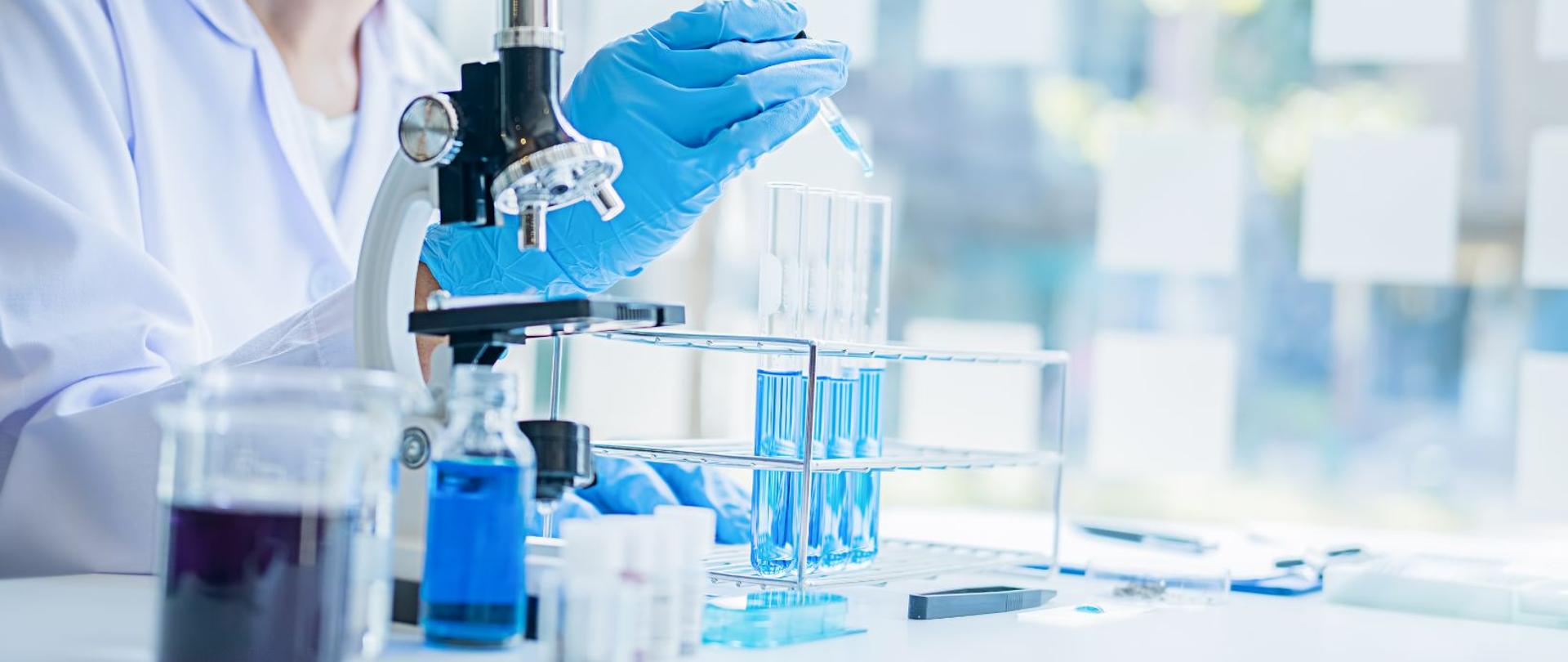 Osoba na zdjęciu trzyma probówkę z niebieskim płynem, obok stoi stojak na probówki oraz mikroskop