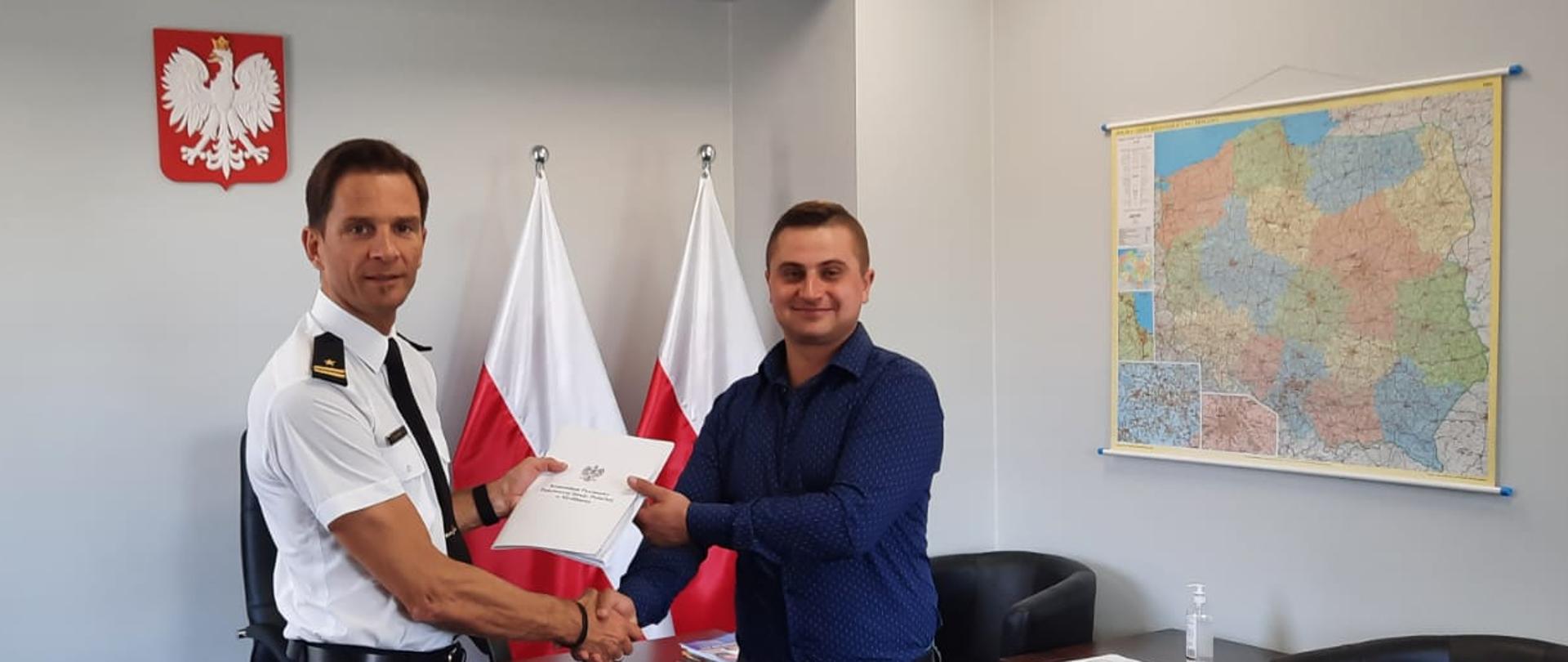 Na zdjęciu widzimy dwóch mężczyzn, którzy podają sobie ręce w uścisku oraz przekazują białą teczkę. Mężczyźni stoją w pomieszczeniu, na ścianie znajduje się mapa Polski i Godło Państwowe.