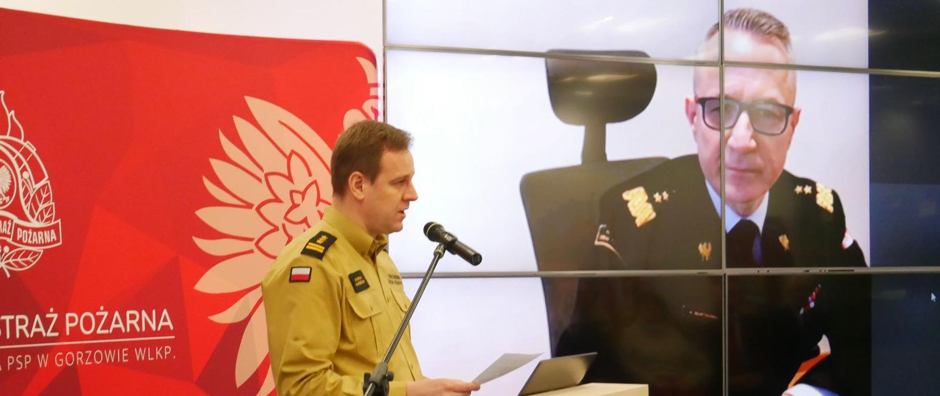 Trwa narada roczna, po lewo przy mikrofonie w sali przemawia komendant wojewódzki, po prawej stronie na telebimie widoczny komendant główny PSP, uczestniczący online w naradzie