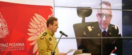Trwa narada roczna, po lewo przy mikrofonie w sali przemawia komendant wojewódzki, po prawej stronie na telebimie widoczny komendant główny PSP, uczestniczący online w naradzie
