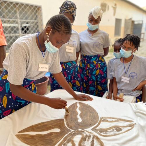 Grupa młodych kobiet stoi wokół stołu, na którym jedna z nich tworzy rysunek na tkaninie