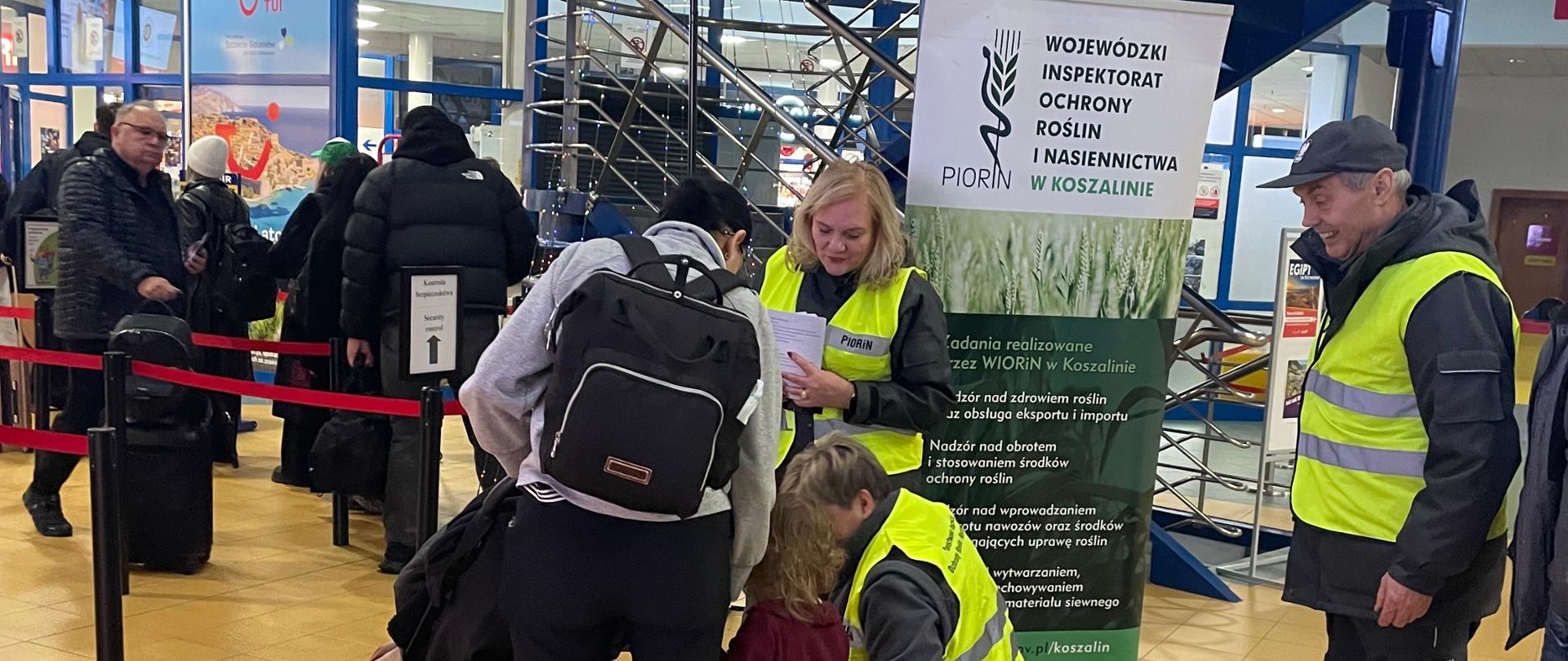 Zdjęcie przedstawia pracowników WIORiN w Porcie Lotniczym Szczecin–Goleniów rozmawiających z pasażerami czekającymi na swój lot. Pracownik WIORiN wręcza dziecku ulotkę oraz materiały promocyjne.