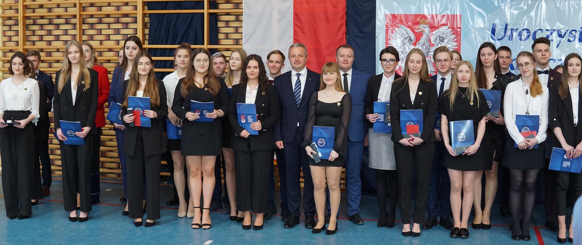 
Grupa ludzi w galowych strojach podczas uroczystości w sali gimnastycznej
