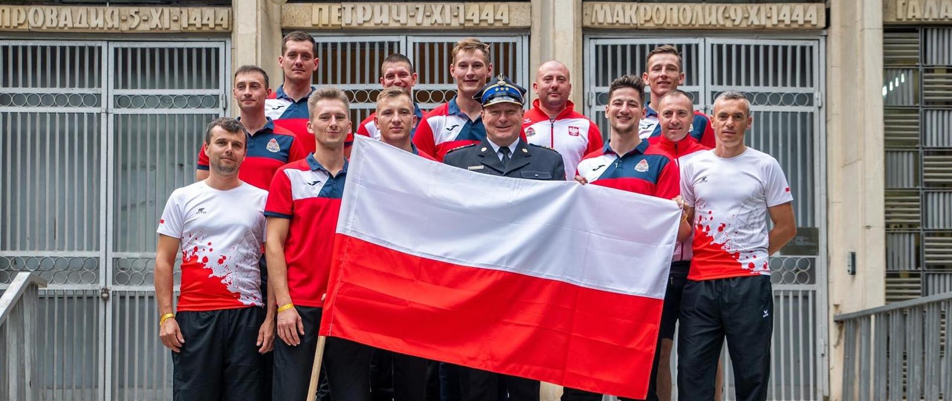 Polska reprezentacja pozuje do zdjęcia grupowego trzymając flagę Polski, w tle budynek