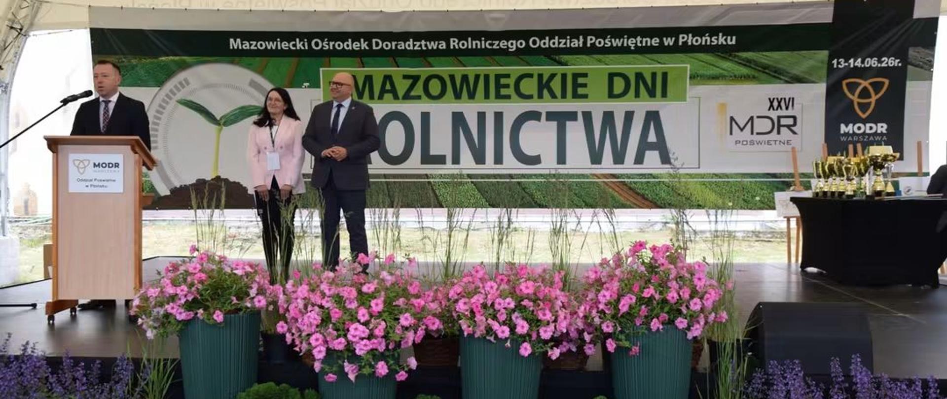 Na scenie przy mównicy z mikrofonem mężczyzna. Obok kobieta i mężczyzna. Wszyscy w garniturach. W tle baner Mazowieckie Dni Rolnictwa. Scena z przodu udekorowana kolorowymi kwiatami i roślinami. Po prawej stronie stół z pucharami.