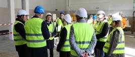 Grupa osób ubranych w kaski ochronne i kamizelki odblaskowe stoi w hali przemysłowej podczas wizyty na terenie budowy lub modernizowanego obiektu. Uczestnicy zgromadzeni są w kręgu i słuchają informacji dotyczących inwestycji.