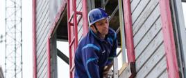 Na zdjęciu z bliższego ujęcia widać strażaka w ubraniu sportowym, który wspina się po drabinie hakowej po piętrach wspinalni 3 piętrowej. W tle jednostka ratowniczo-gaśnicza JRG Nr 1 w Ostrowcu Św.