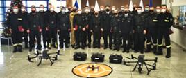 Zdjęcie grupowe uczestników szkolenia. Na pierwszym planie 3 drony w tle strażacy stojący w dwuszeregu w mundurach w kolorze czarnym. 
