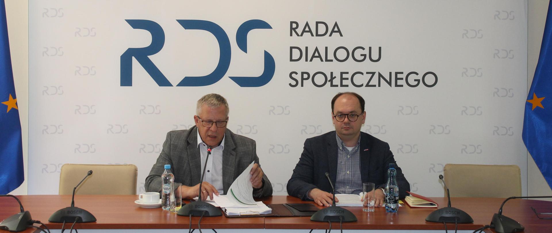 Na zdjęciu widnieje stół prezydialny za którym siedzi Przewodniczący Zespołu ds. budżetu, wynagrodzeń i świadczeń socjalnych – Leszek Miętek a po Jego lewej Sebastian Gajewski – Podsekretarz Stanu w Ministerstwie Rodziny, Pracy i Polityki Społecznej. Po obu stronach stołu widnieją flagi UE. W tle siedzących znajduje się baner z napisem Rada Dialogu Społecznego a nad nim wisi godło Polski.