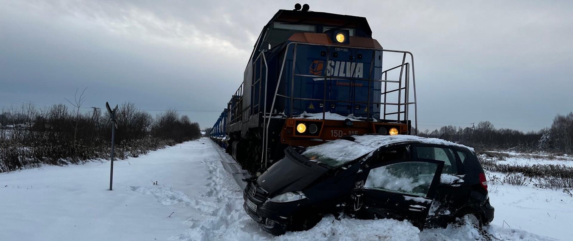 W warunkach zimowych, na torowisku widoczny samochód osobowy, który zderzył się z lokomotywą. Lokomotywa przylega bezpośrednio do pojazdu. 