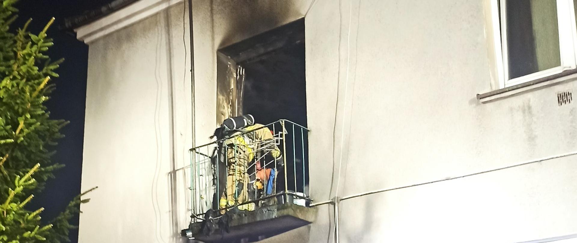 Zdjęcie obrazuje strażaka pracującego na balkonie budynku wielokondygnacyjnego na ścianie widoczne oznaki pożaru
