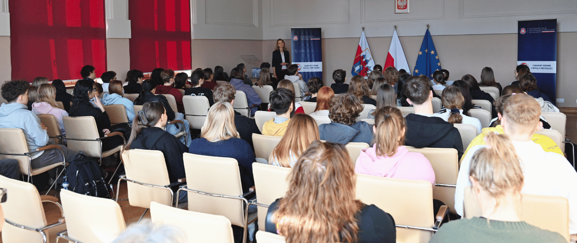 Uczniowie siedzący na sali konferencyjnej słuchają prelekcji prowadzonej przez kobietę stojącą z przodu, obok flag Polski i Unii Europejskiej oraz banerów informacyjnych.