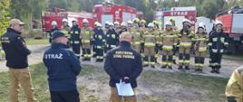 Grupa osób ubranych w ubrania specjalne straży pożarnej stoi w dwuszeregu, przed nimi stoja mężczyżni w mundurach służbowych straży pożarnej, za nimi widać wozy gaśnicze