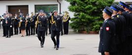 Na placu przed budynkiem odbywa się uroczysty apel strażacki. Mazowiecki Komendant Wojewódzki PSP i Dowódca uroczystości salutuje w kierunku ustawionego w szeregu pododdziału. W tle widoczna jest orkiestra dęta w galowych mundurach, a obok rośnie duże drzewo iglaste. Atmosfera wskazuje na oficjalne obchody
