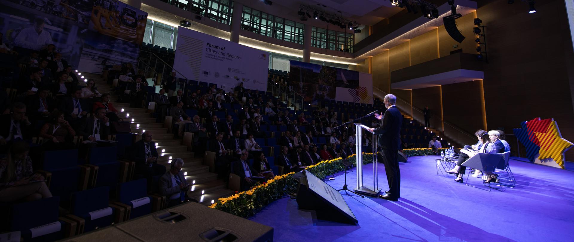 Zdjęcie robione od strony sceny, na środku sceny minister Jerzy Kwieciński przy mównicy, po jego prawej stronie pięcioro prelegentów na fotelach oraz stojące logo wydarzenia. W tle nieoświetlona widownia, ścianki promocyjne z logotypami i nazwą "Forum of Cities and Regions"