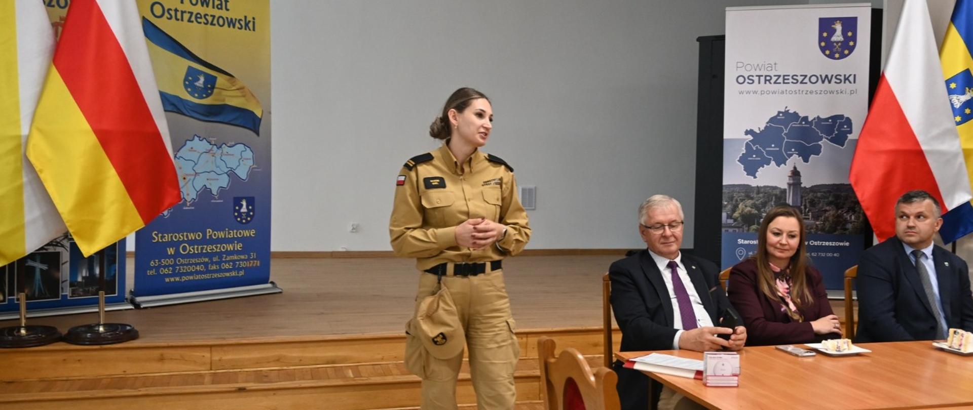 Strażak w trakcie prezentacji. Po prawej stronie siedzą słuchacze, w tle flagi i banery powiatu ostrzeszowskiego.