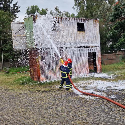 Dwie osoby w strojach strażackich oblewają wodą z węża metalowy kontener