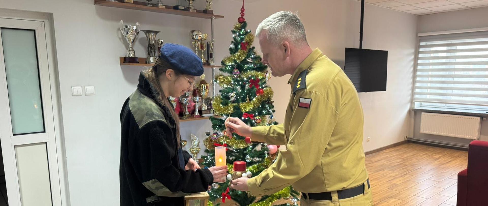 Młoda osoba w granatowym berecie i ciemnej kurtce trzyma zapaloną świecę w lampionie, a obok stoi mężczyzna w mundurze, który pomaga ją zapalić; w tle widać udekorowaną choinkę i półki z pucharami.