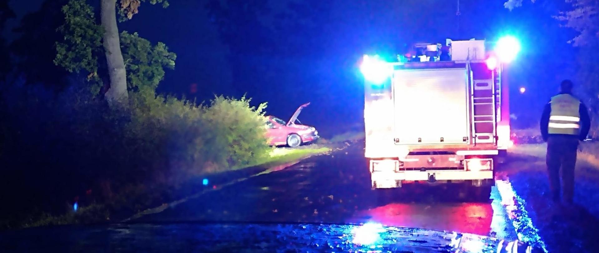 Mokra nawierzchnia drogi. Pora nocna. Teren oświetlają halogeny pojazdów pożarniczych zaparkowanych na drodze oraz niebieskie światła błyskowe. Na poboczu drogi Policjant w kamizelce odblaskowej prowadzi czynności. Czerwony samochód uczestniczący w zdarzeniu stoi na skrzyżowaniu. Ma podniesioną maskę przednią.