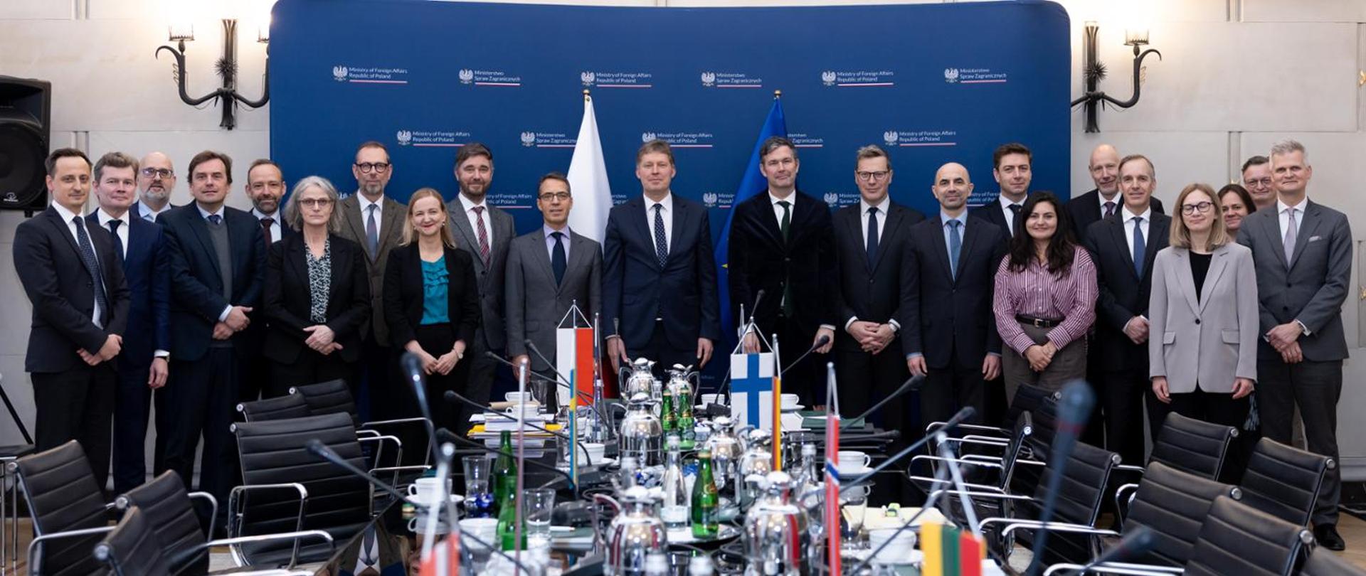 A group of participants stands in a conference room during an official meeting. A conference table with microphones and national flags is visible in the foreground, with a backdrop displaying the logo of the Polish Ministry of Foreign Affairs in the background.
