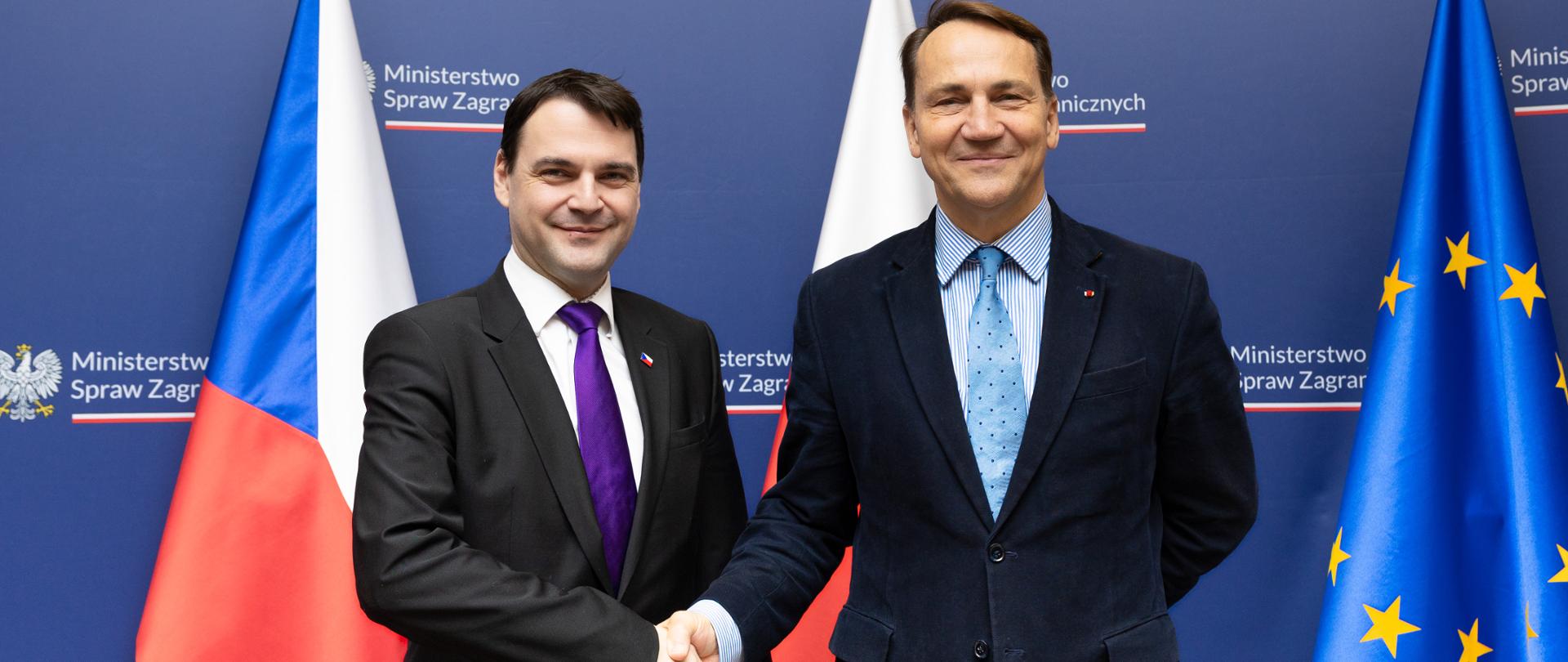 Dwóch mężczyzn w garniturach podczas uścisku dłoni. W tle niebieska ścianka oraz flagi. 