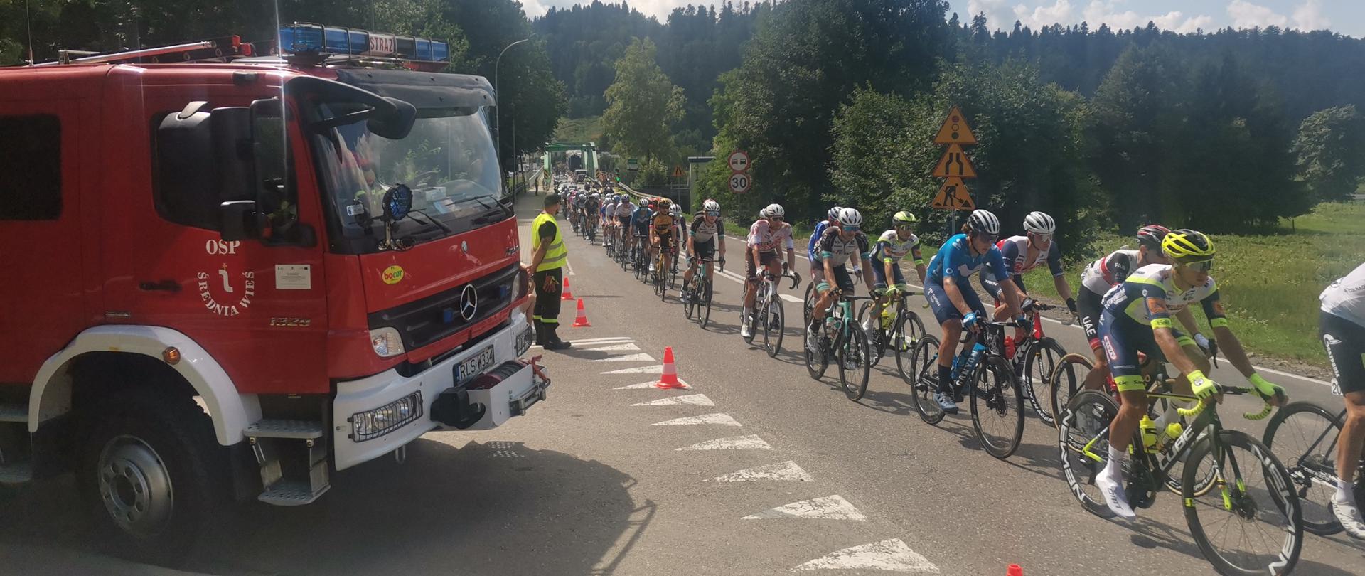 Na zdjęciu widzimy grupę kolarzy , samochód Ratowniczo-Gaśniczy Ochotniczej Straży Pożarnej w Średniej Wsi oraz strażaka który zabezpiecza wyznaczony odcinek drogi.