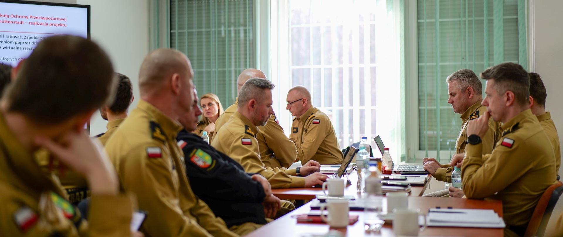 Sala konferencyjna wypełniona strażakami PSP w mundurach piaskowych. Uczestnicy siedzą przy długim stole, mają przed sobą laptopy, notatniki i kubki z kawą. W tle na ekranie wyświetlony jest slajd prezentacji na temat międzynarodowej współpracy strażackiej.