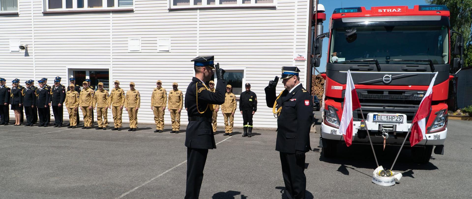 Na zdjęciu widać Dowódcę uroczystości salutującego przed Komendantem Wojewódzkim podczas składania meldunku. W tyle z prawej strony widać funkcjonariuszy KP PSP w Wieruszowie stojących w rzędzie w umundurowaniu na tle budynku JRG. Z lewej strony zdjęcia widać samochód pożarniczy, czerwony przed którym postawione są dwie flagi biało-czerwone.