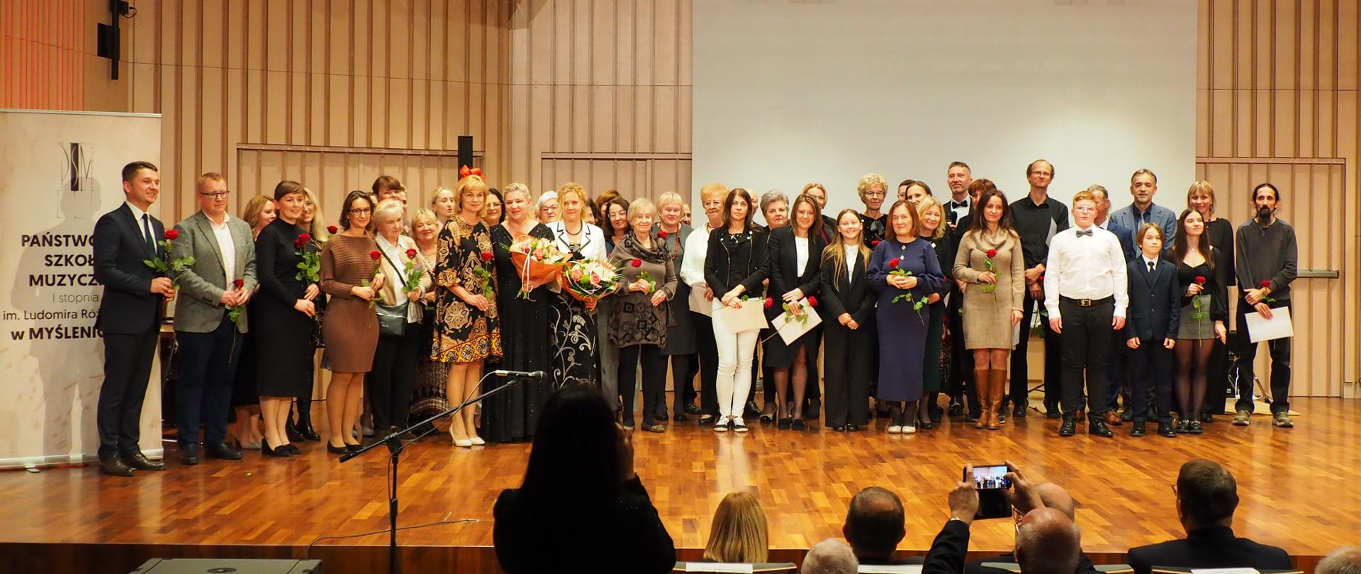 Grupa osób z kwiatami stoi na scenie w sali koncertowej, po lewej stronie plakat z logo szkoły i napisem Państwowa Szkoła Muzyczna I stopnia im. Ludomira Różyckiego w Myślenicach.