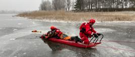 Zdjęcie przedstawia strażaków ćwiczących na lodzie. Strażacy zabezpieczeni w ubrania do pracy w wodzie. Jeden ze strażaków topi się w przeręblu. Dwóch strażaków zabezpieczonych liną, w tym jeden leży na desce lodowej i ewakuuje z wody osobę topiącą się.