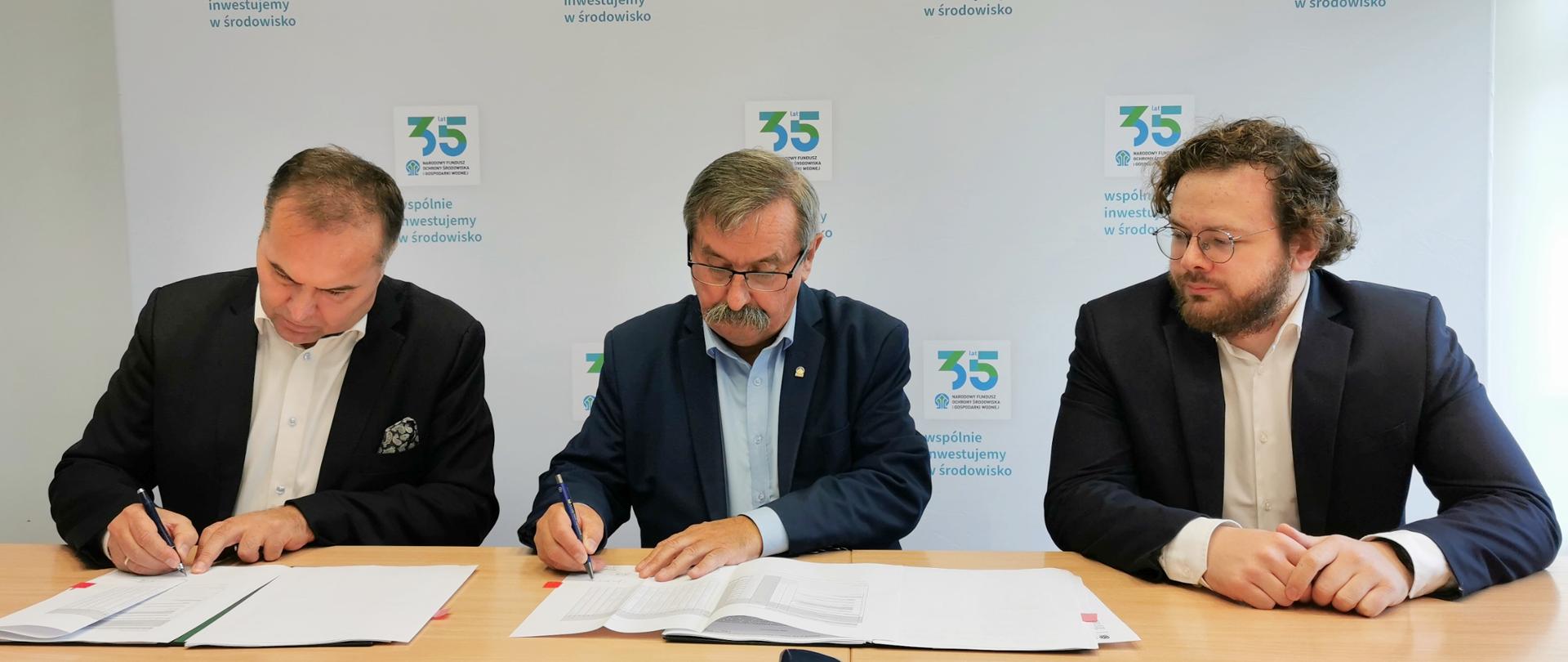 1. Zdjęcie przedstawia uroczystość podpisania umowy na dofinansowanie projektu Konwersja kotła węglowego na kocioł opalany biomasą wraz z układem gospodarki paliwem biomasowym pomiędzy NFOŚiGW a QEMETICA Soda Polska S.A.. Na zdjęciu trzech mężczyzn siedzących przy stole, podpisują dokumenty. W tle roll-up z logotypami NFOŚiGW. Na zdjęciu od lewej: Tomasz Molenda, Prezes Zarządu QEMETICA Soda Polska S.A., Józef Matysiak, Zastępca Prezesa Zarządu NFOŚiGW, Piotr Kapuściński, Wiceprezes Zarządu QEMETICA Soda Polska S.A.