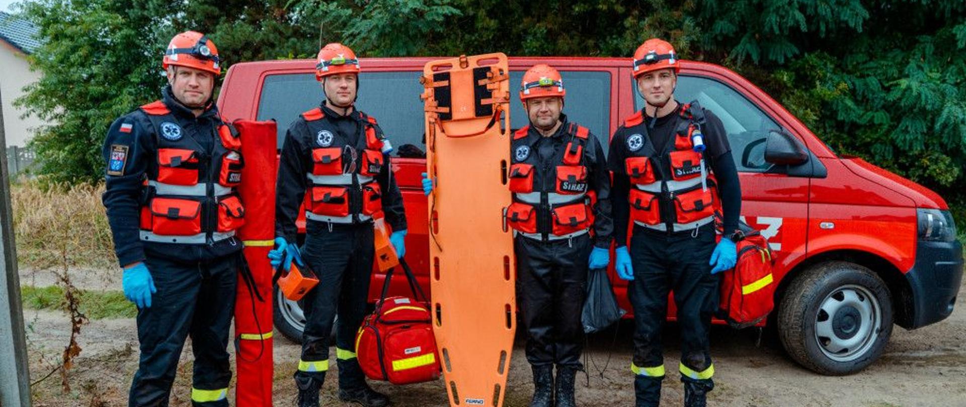 „Czterech strażaków w kaskach i kamizelkach ratowniczych stoi przed czerwonym samochodem straży pożarnej. Trzymają sprzęt ratowniczy, w tym nosze, torby medyczne i rulon zabezpieczeniowy.”