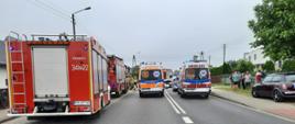Na zdjęciu widać samochody gaśnicze z JRG 1 i 2. Pojazdy są w kolorze czerwonym, posiadają sygnalizację ostrzegawczą. Obok karetki Państwowego Ratownictwa Medycznego. Niebo zachmurzone. Po prawej stronie osoby postronne. Za pojazdami funkcjonariusze Policji oraz straży pożarnej.
