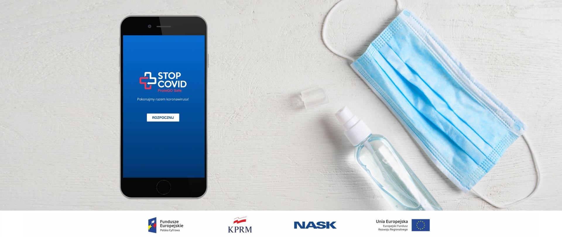 Z prawej strony - leżące na białym tle maseczka chirurgiczna (widoczna niebieską stroną) i przezroczyste opakowanie z płynem do dezynfekcji. Po lewej stronie czarny telefon z ekranem startowym aplikacji STOP COVID - ProteGO Safe.