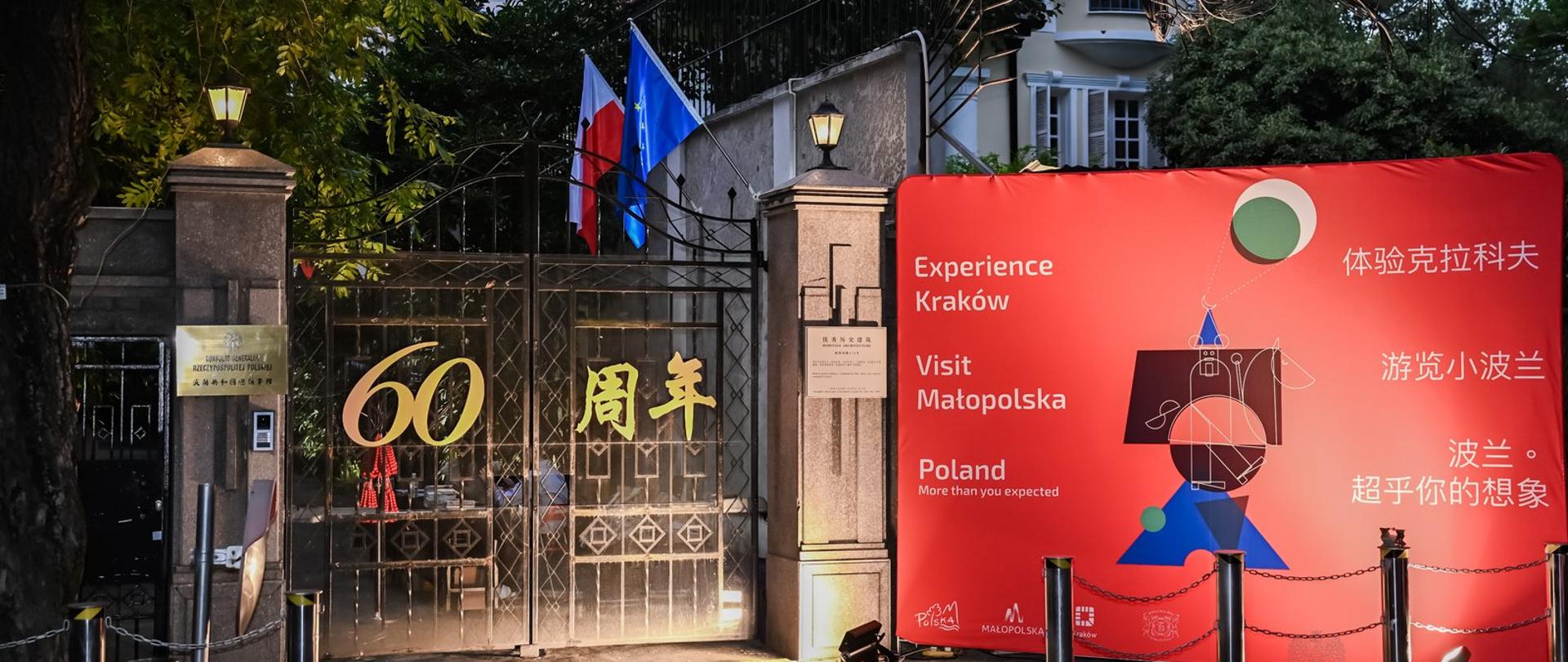 Brama konsulaty ze złotym napisem 60-lat, obok czerwony baner promujący Kraków, Małopolskę i Polskę.