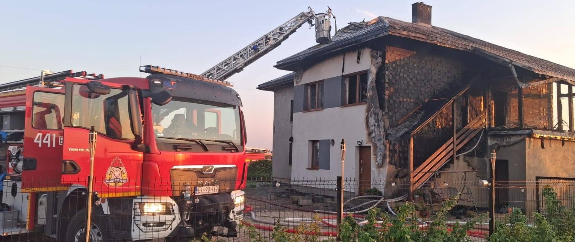 Na zdjęciu widoczny jest front budynku oraz prowadzone działania gaśnicze z wykorzystaniem drabiny mechanicznej. Na pierwszym planie znajduje się samochód ratowniczo-gaśniczy. Na budynku widoczne są rozległe uszkodzenia elewacji, balkonu i schodów zewnętrznych.