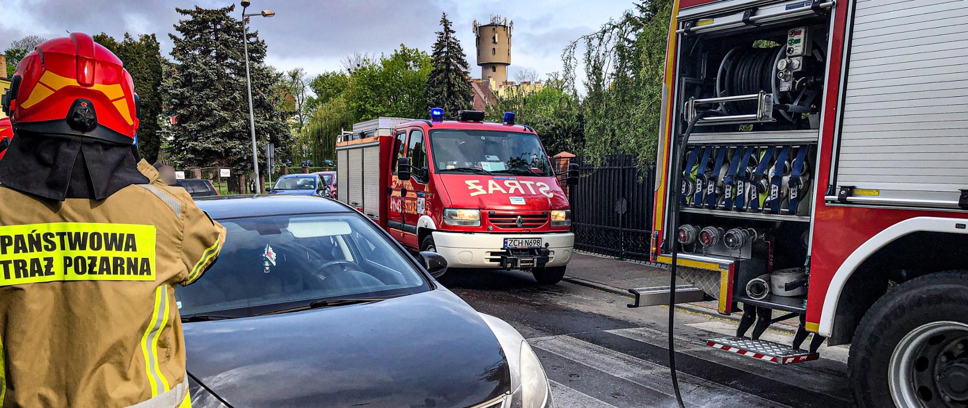 W centrum fotografii znajduje się czarny samochód osobowy. Dookoła niego na asfalcie jest rozsypany proszek z gaśnicy. Po lewej stronie stoi ubrany w ubranie specjalne strażak. A po prawej stronie znajdują się dwa wozy strażackie, 