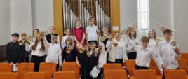 Na zdjęciu widoczna jest grupa uczniów klasy trąbki pozujących w sali koncertowej. Dzieci i młodzież stoją między rzędami pomarańczowych krzeseł, trzymając w rękach trąbki, część z nich unosi instrumenty w górze. Uczniowie ubrani są w stroje koncertowe w jasnych i ciemnych kolorach. W tle znajduje się prospekt organowy z piszczałkami oraz jasne ściany sali. Zdjęcie przedstawia radosny moment po wspólnym występie muzycznym.