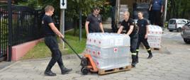 Czterech strażaków transportuje na tzw. "paleciaku" pojemniki plastikowe z płynem dezynfekcyjnym. Pojemniki ułożone są w trzech warstwach. Każda z warstw jest ostreczowana. W tle naczepa ciągnika siodłowego, którym przywiezione zostały środki.