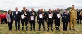 Zdjęcie przedstawia grupę dziewięciu mężczyzn i jednej kobiety, ustawionych w rzędzie na trawiastym polu. Osoby po lewej i prawej stronie rzędu, a także w jego środku, mają na sobie mundur strażacki w czarnym kolorze, a na ich głowach widać szare lub białe nakrycia głowy. Mężczyzna po prawej stronie jest ubrany w beżowy mundur. Mężczyzna w środku rzędu i po lewej stronie, ubrany w ciemną marynarkę i niebieskie spodnie, ma w rękach nagrody. Na trawiastym polu widoczne są ścieżki i inne osoby w tle, a za nimi znajduje się zielony teren z domami. Niebo jest pochmurne, ale jasne.
