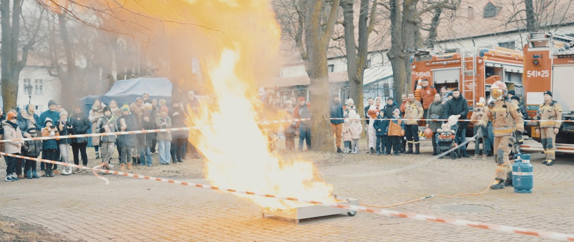 Na zdjęciu moment intensywnego spalania oleju po wlaniu do niego wody. Duży słup ognia. Na około za taśmami ostrzegawczymi widzowie i wozy strażackie.