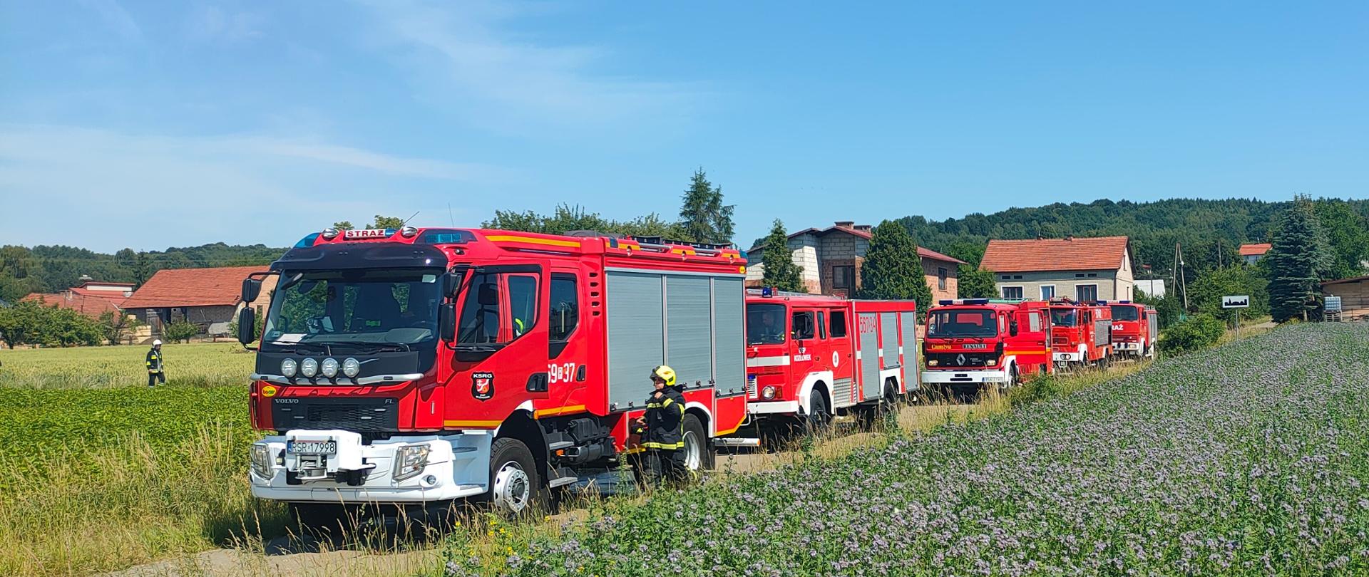 na zdjęciu widoczne 5 samochodów ratowniczo-gaśniczych zaparkowane jeden za drugim na drodze . Dookoła widoczne łąki w tle budynki jednorodzinne oraz las.