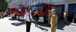 Strażak ubrany w mundur wyjściowy melduje komendantowi głównemu Państwowej Straży Pożarnej za nimi przed bramami stoją samochody ratowniczo-gaśnicze.