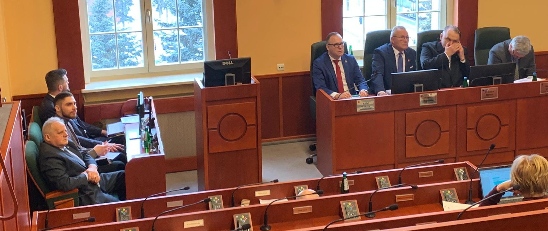 grupa ludzi w garniturach, w tym wicewojewoda, siedzą przy biurkach. W tle wyświetla się na ekranie tytuł wydarzenia na którym się zgromadzili. 