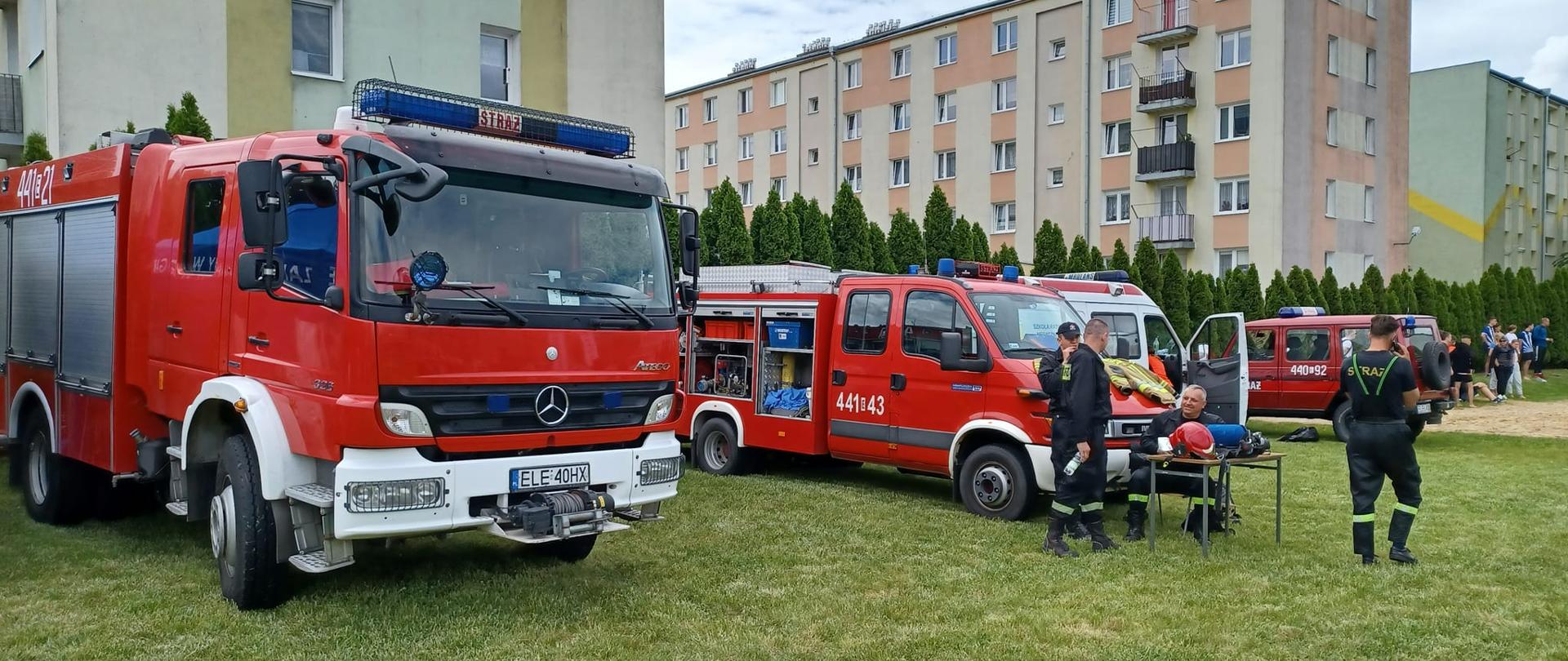 Na zdjęciu widać czerwone samochody strażackie, karetkę pogotowia oraz strażaków w czarnych strojach. Jeden samochód strażacki ma otwarte boczne skrytki, w których znajduje się sprzęt ratowniczy. Za strażakami widać kolorowe bloki oraz tuje. W oddali widać osoby biorące udział w pikniku. Wszyscy stoją na trawie. Jest widno.