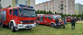 Na zdjęciu widać czerwone samochody strażackie, karetkę pogotowia oraz strażaków w czarnych strojach. Jeden samochód strażacki ma otwarte boczne skrytki, w których znajduje się sprzęt ratowniczy. Za strażakami widać kolorowe bloki oraz tuje. W oddali widać osoby biorące udział w pikniku. Wszyscy stoją na trawie. Jest widno.
