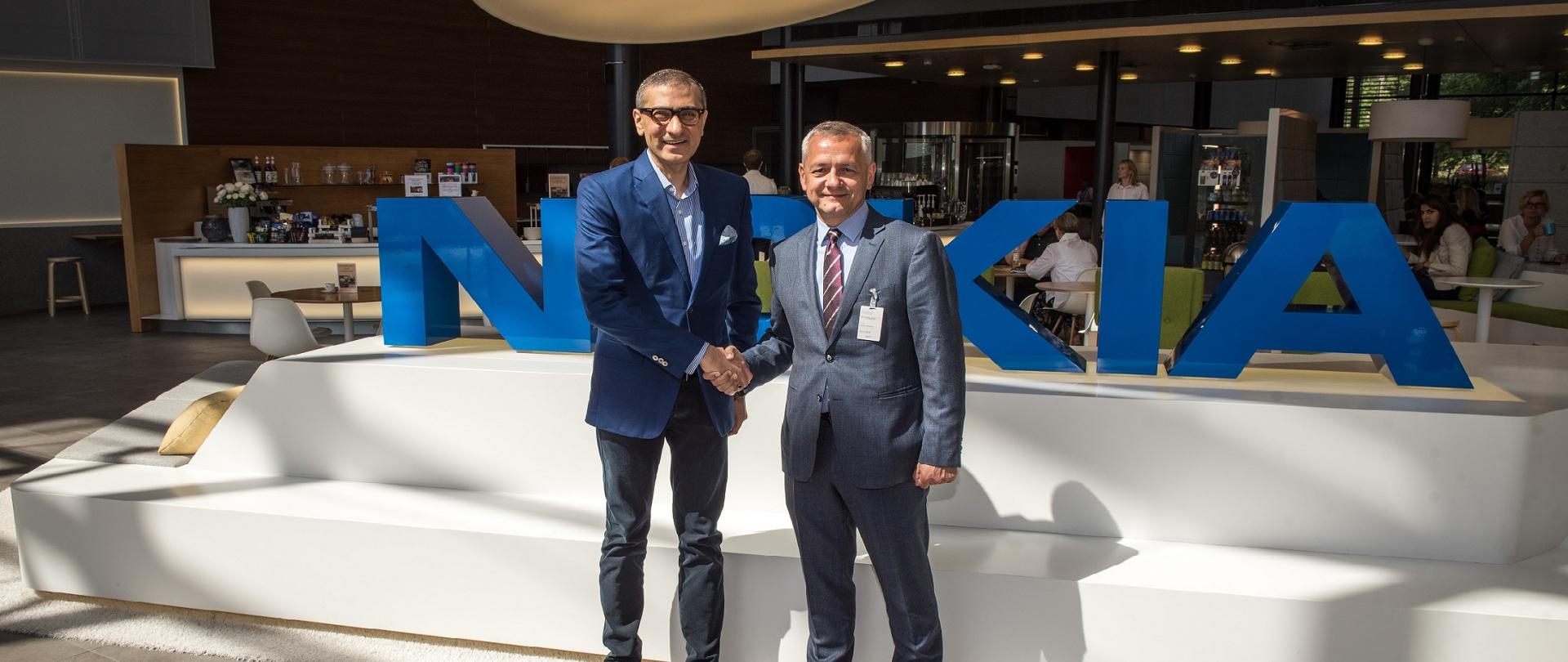 Minister cyfryzacji i CEO Nokii pozują do zdjęcia podając sobie ręce. W tle logo Nokii, dalej część restauracyjna firmy, w której przy stolikach siedzi kilka osób.