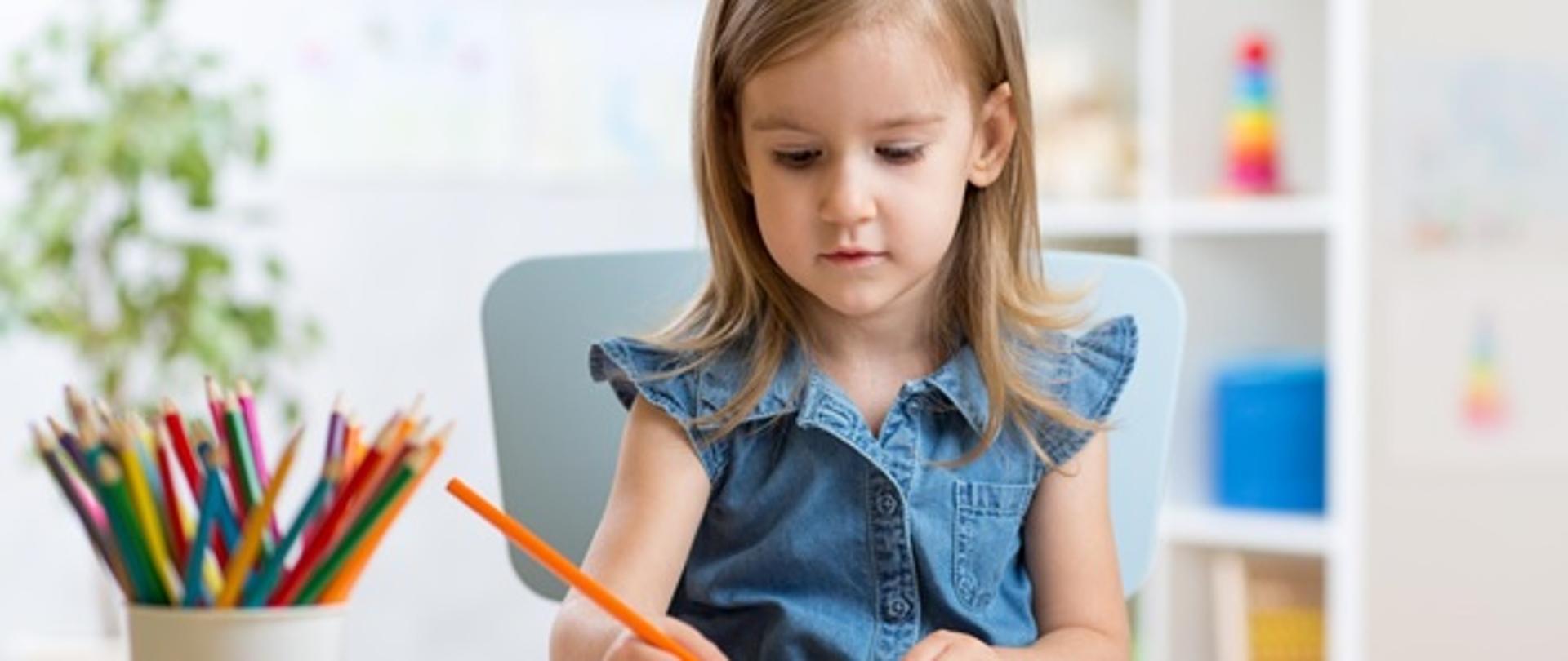 dziewczynka siedząca w fotelu przy stole rysująca kredkami. Na stole znajduje się kubek z kredkami